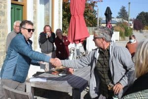 Two men shaking hands outdoors with people clapping nearby