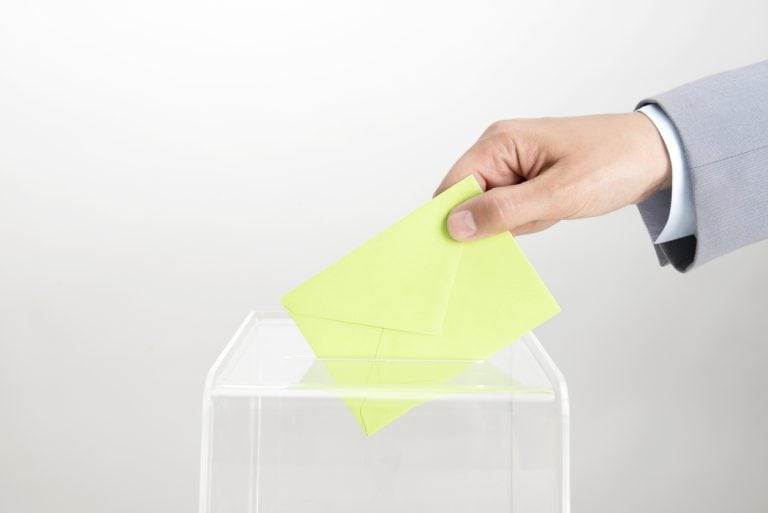 Human hand is inserting green envelope into ballot box.