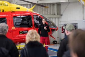 Man speaking with microphone in front of red and yellow air rescue helicopter
