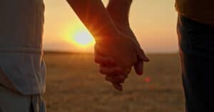 Close-up of two hands holding each other at sunset in an open field, symbolizing connection, love, and support. The golden light of the setting sun creates a warm and intimate atmosphere.