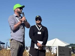 Two men speaking at outdoor agricultural event with tents and crops.